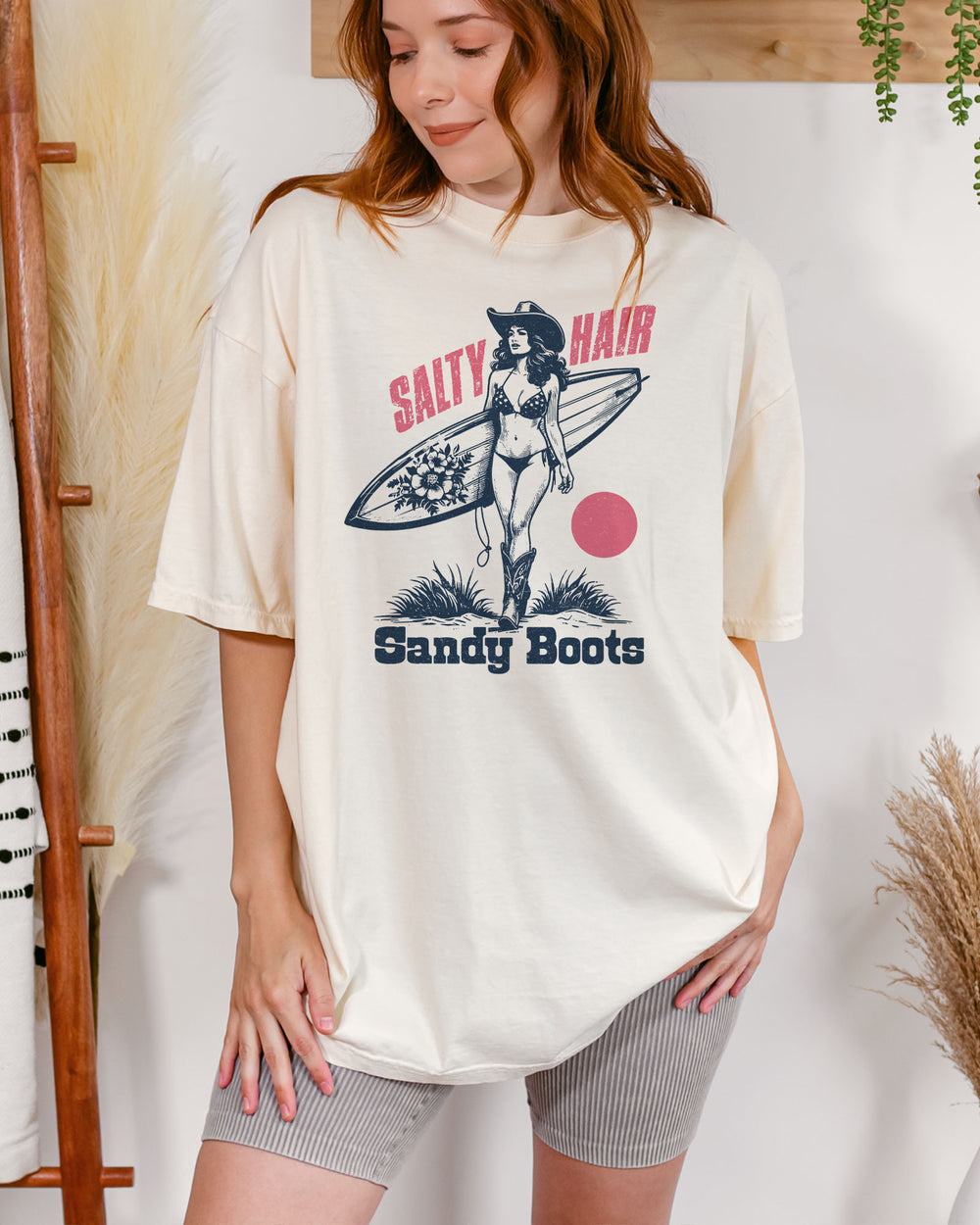 Woman wearing "Salty Hair Sandy Boots" shirt in ivory color with retro graphic of a cowgirl with a surfboard under her arm.
