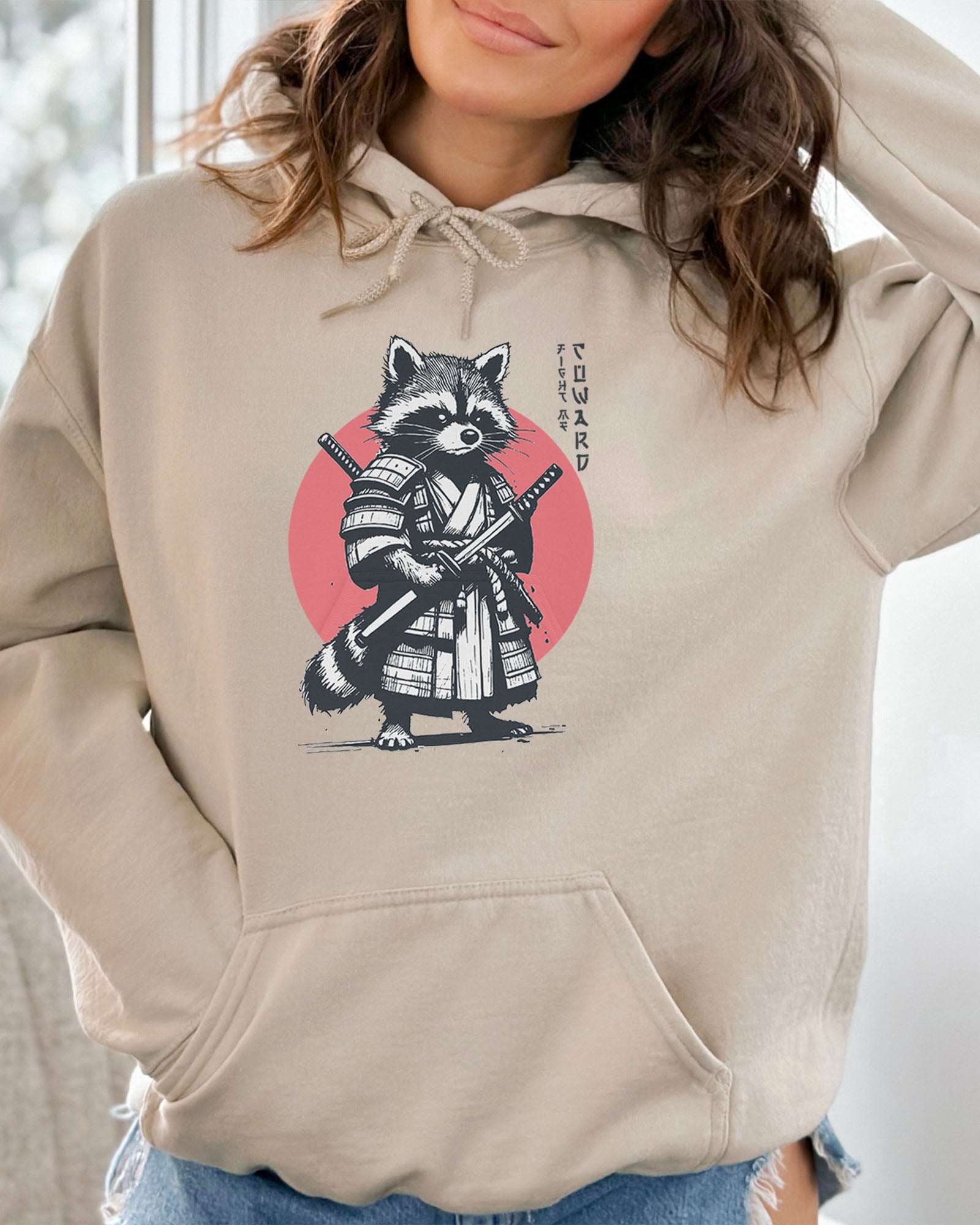 Woman wearing a sand beige hoodie featuring a retro funny samurai raccoon graphic with the quote 'Fight Me Coward' in a Japanese-style font.