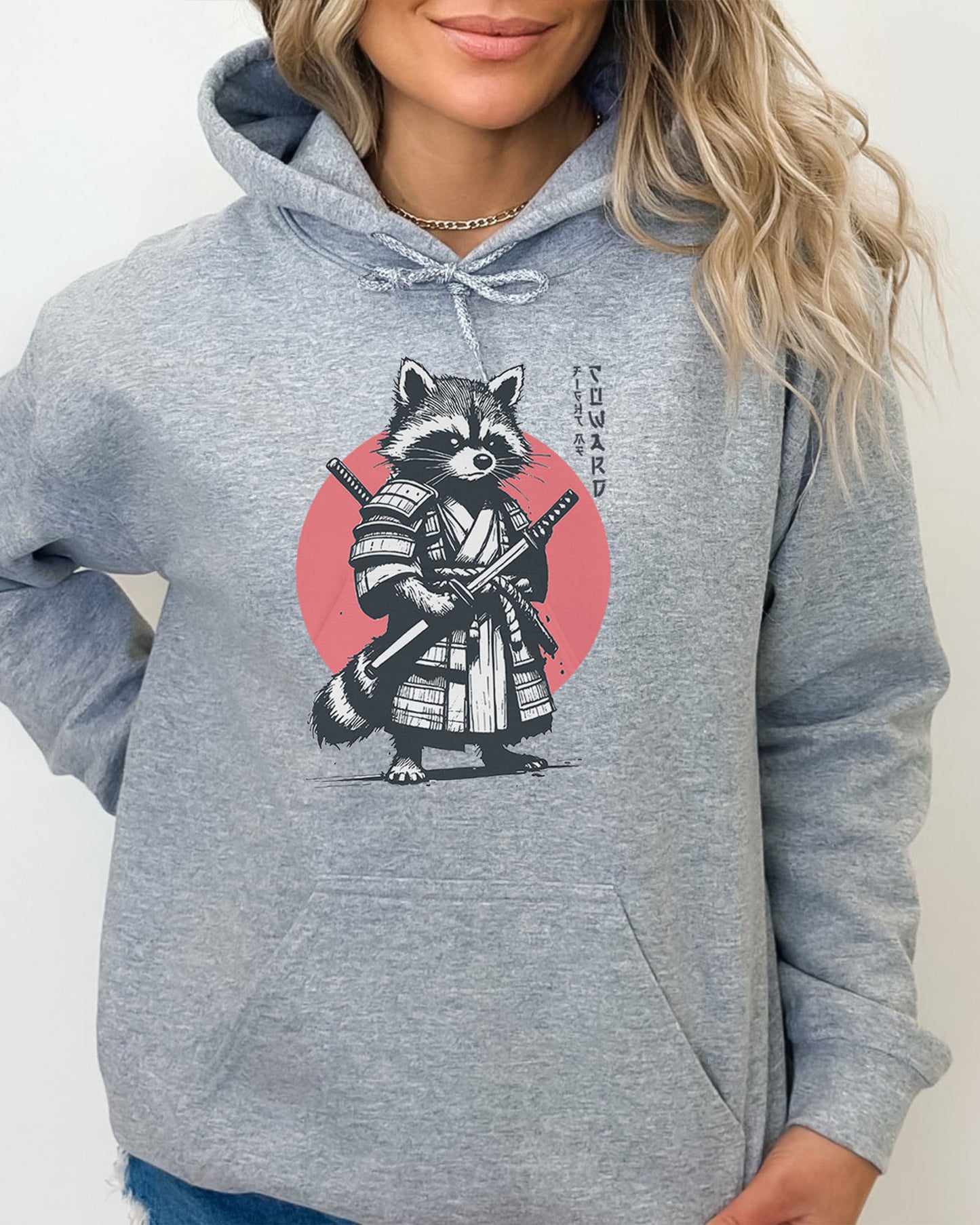 Woman wearing a sport gray hoodie featuring a retro funny samurai raccoon graphic with the quote 'Fight Me Coward' in a Japanese-style font.