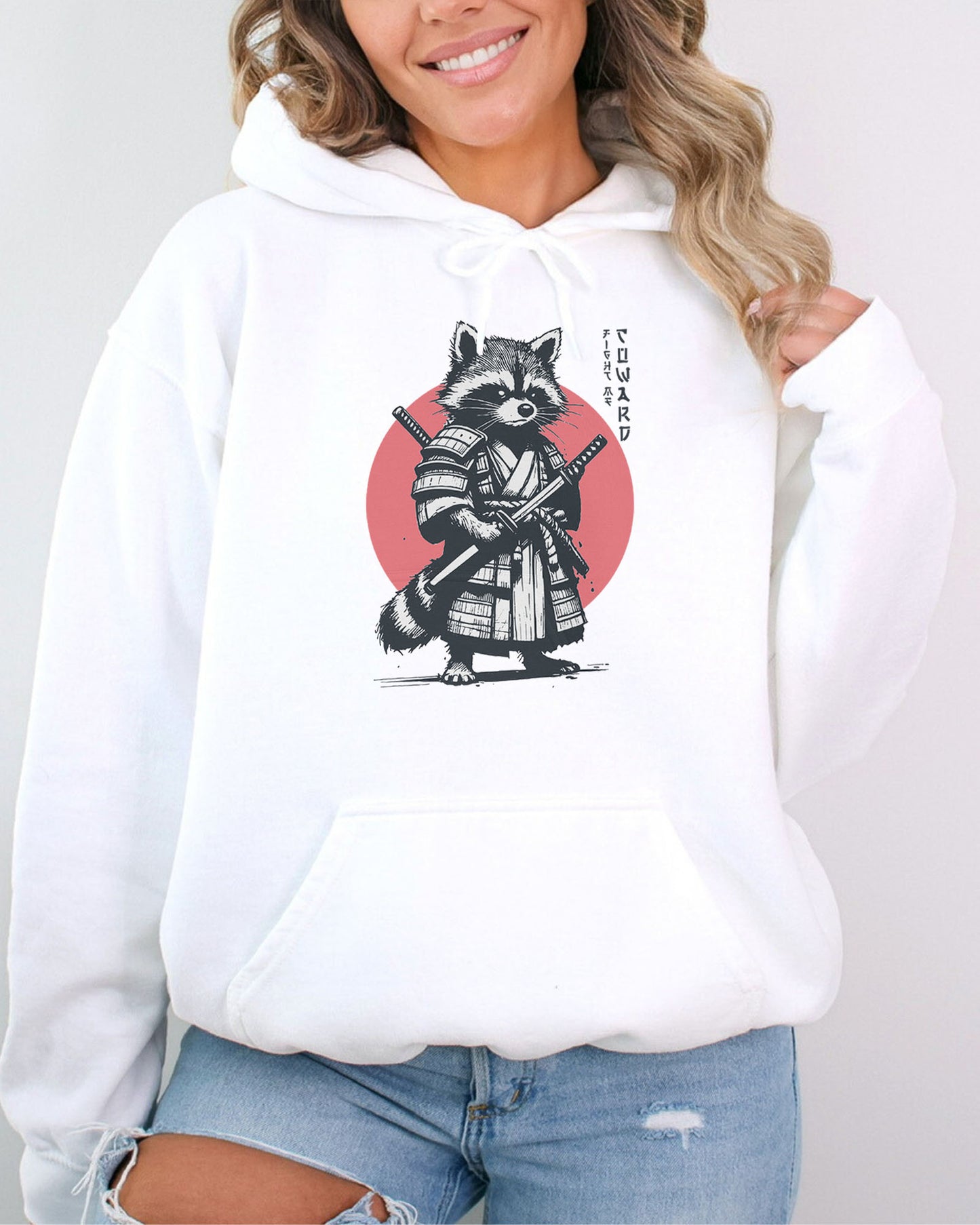 Woman wearing a white hoodie featuring a retro funny samurai raccoon graphic with the quote 'Fight Me Coward' in a Japanese-style font.