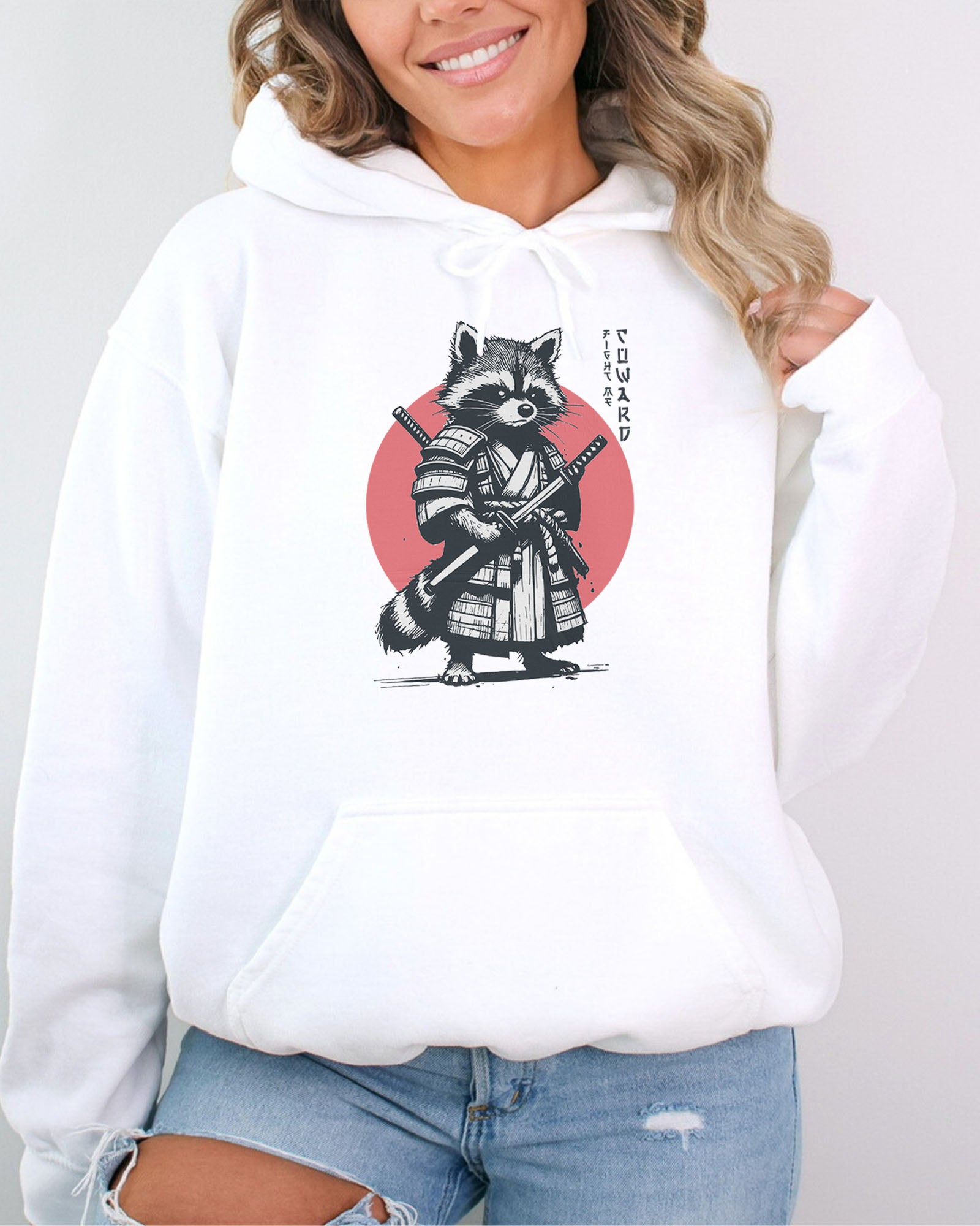 Woman wearing a white hoodie featuring a retro funny samurai raccoon graphic with the quote 'Fight Me Coward' in a Japanese-style font.