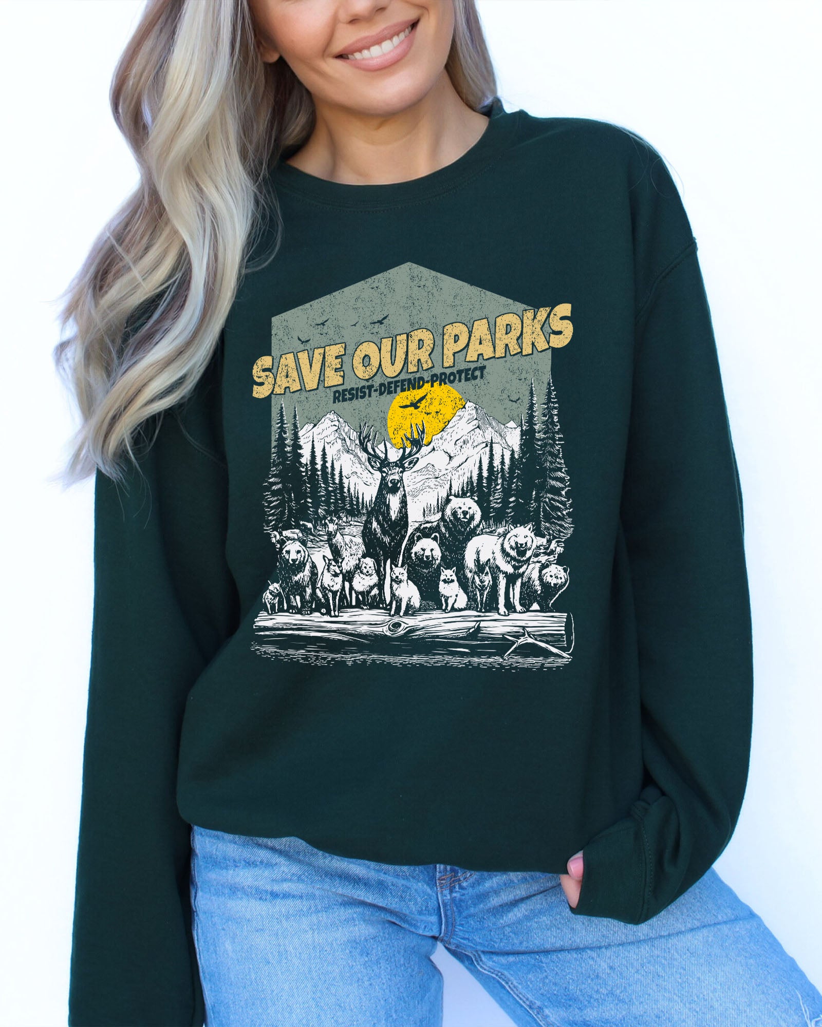 Woman wearing a forest green sweatshirt with a retro wildlife design featuring animals lined up in a park and the quote 'Save Our Parks – Resist, Defend, Protect'.