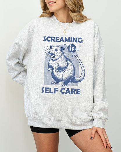 Model wearing ash color sweatshirt with 'Screaming Is Self Care' graphic design
