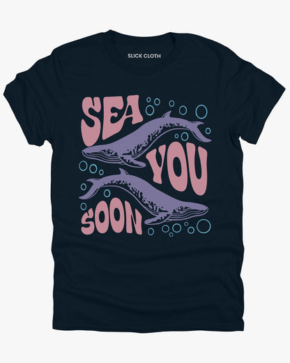 Flat lay of a midnight blue 'Sea You Soon' t-shirt with a retro whales and sea life graphic.