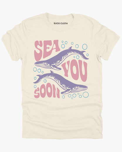 Flat lay of a natural-colored 'Sea You Soon' t-shirt with a retro whales and sea life graphic.