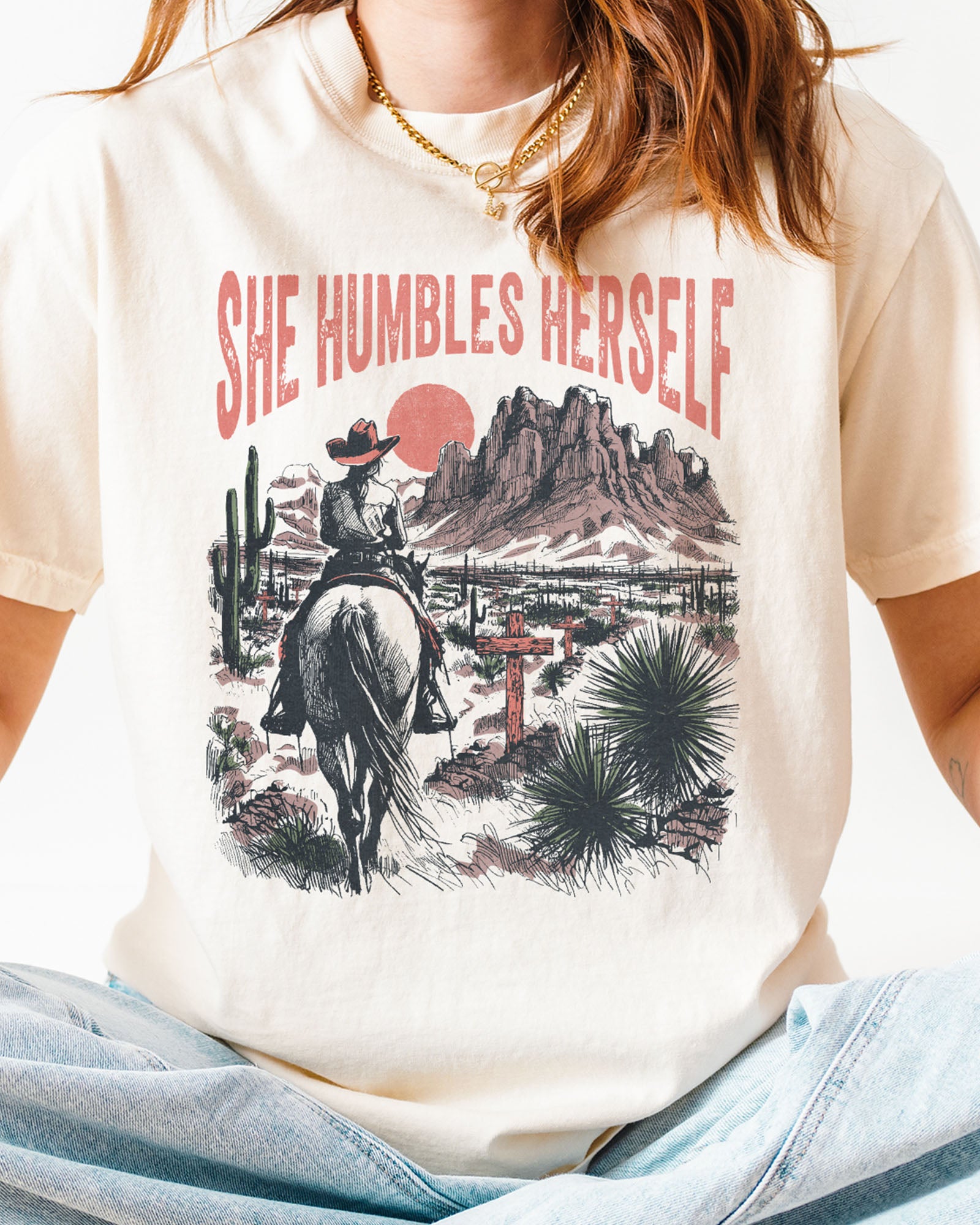 Close-up of woman wearing an ivory "She Humbles Herself" shirt with a retro graphic of a cowgirl riding a horse through the desert.