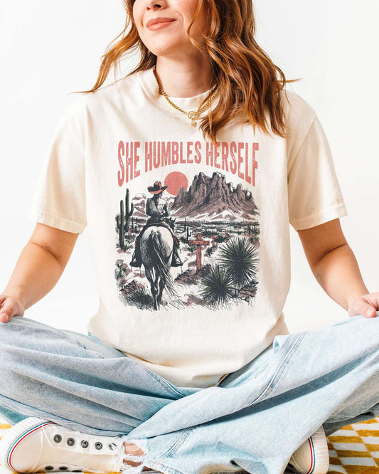 Woman wearing an ivory "She Humbles Herself" shirt with a retro graphic of a cowgirl riding a horse through the desert.