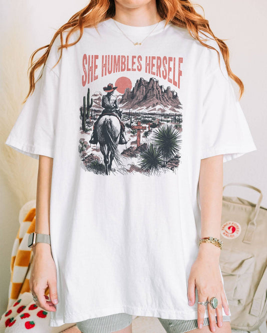 Woman wearing a white "She Humbles Herself" shirt with a retro graphic of a cowgirl riding a horse through the desert.