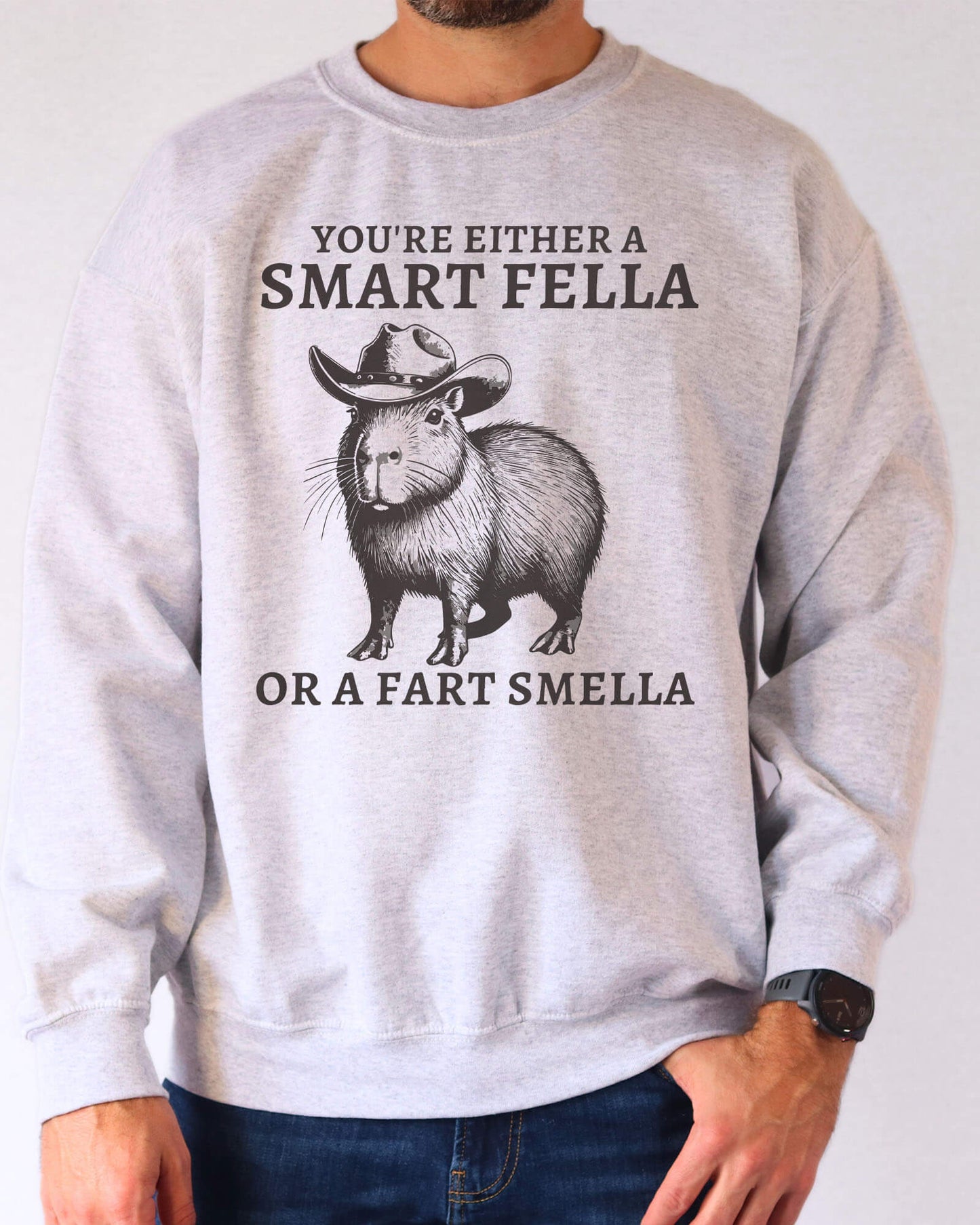 Man wearing an ash gray sweatshirt featuring a funny retro cowboy capybara graphic and the quote 'You're Either A Smart Fella Or A Fart Smella'.
