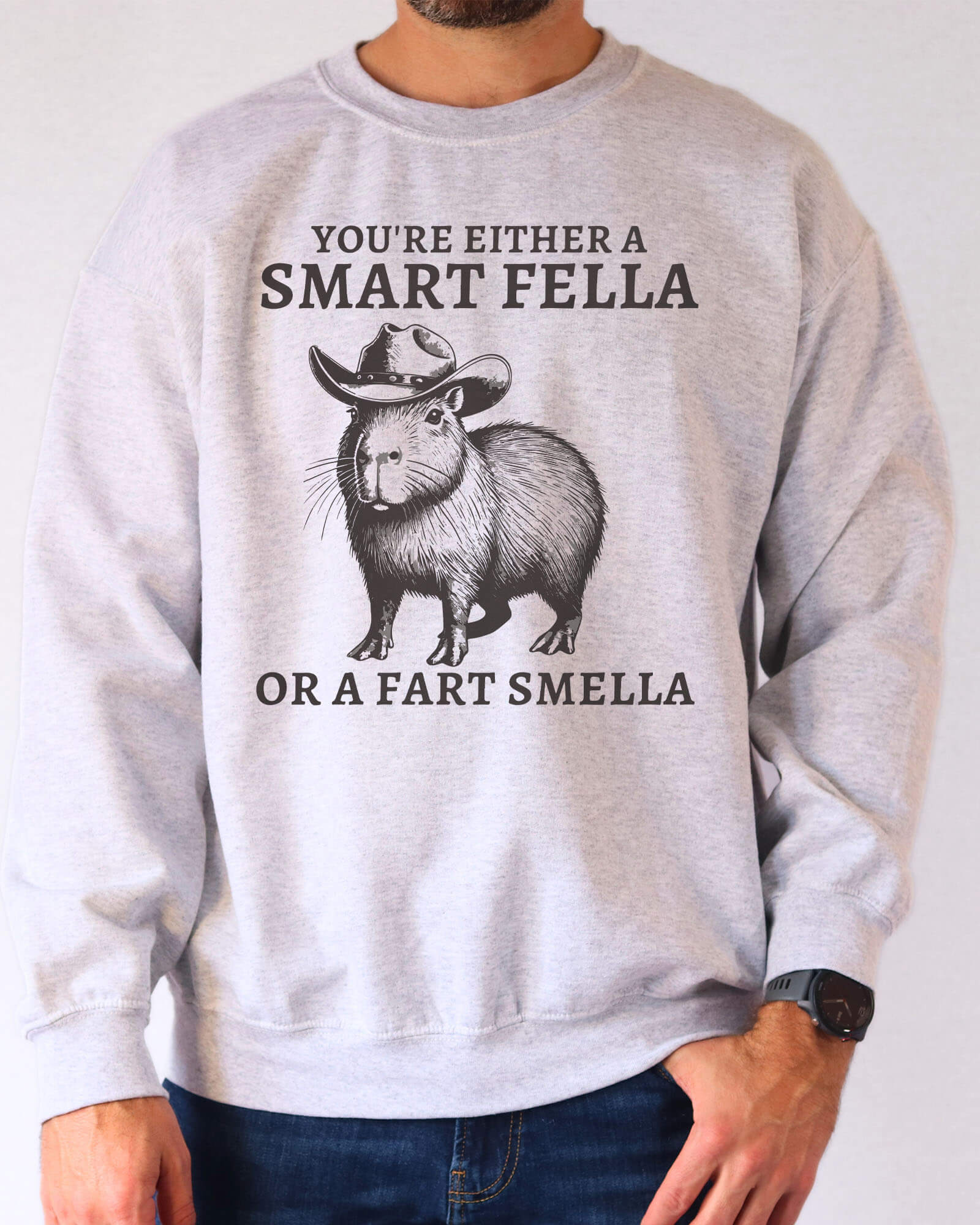 Man wearing an ash gray sweatshirt featuring a funny retro cowboy capybara graphic and the quote 'You're Either A Smart Fella Or A Fart Smella'.