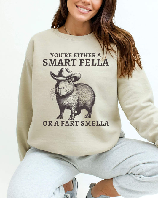 Woman wearing a sand beige sweatshirt featuring a funny retro cowboy capybara graphic and the quote 'You're Either A Smart Fella Or A Fart Smella'.