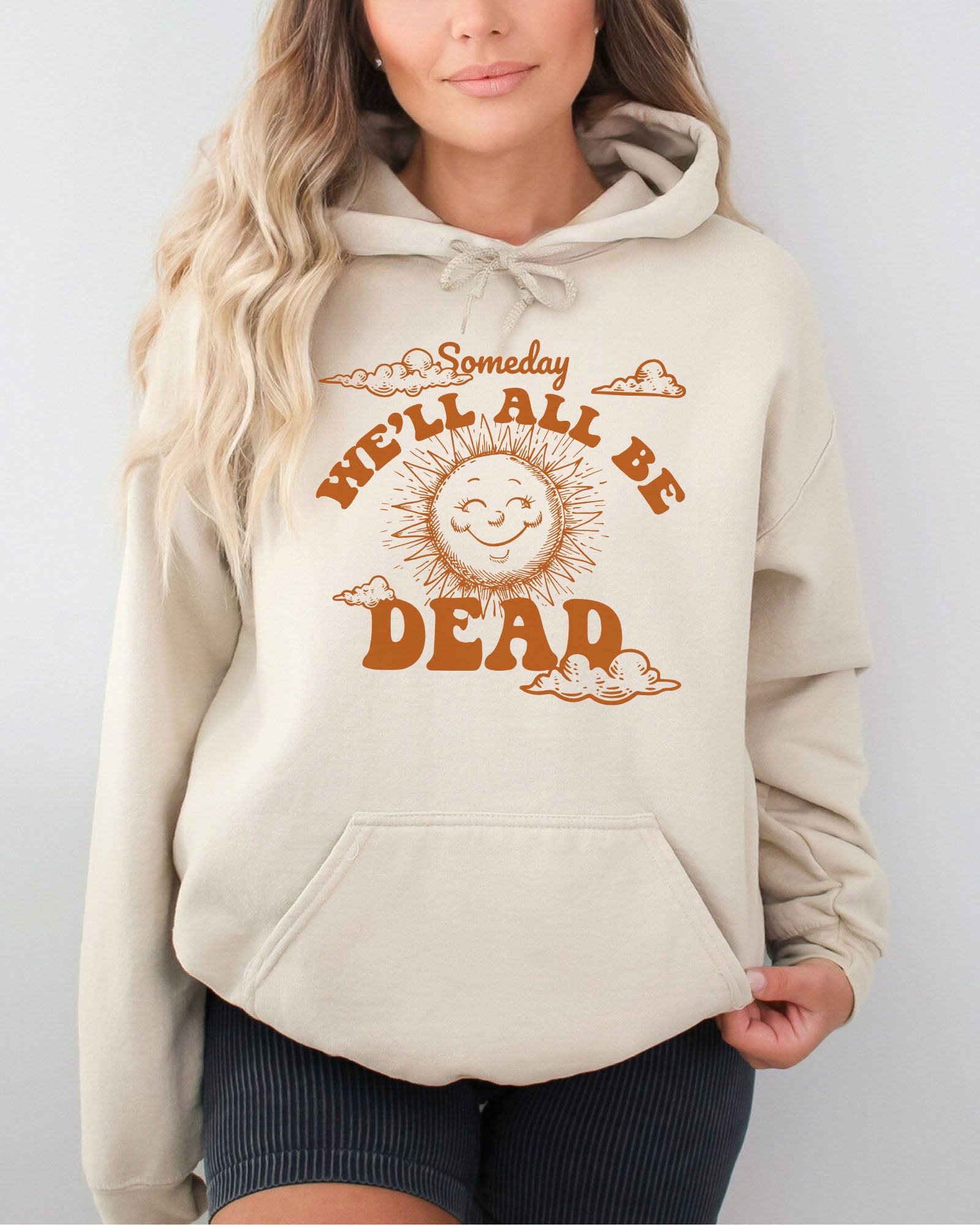 Woman wearing a sand beige hoodie featuring a funny retro-style laughing sun graphic and the quote 'Someday We'll All Be Dead'.