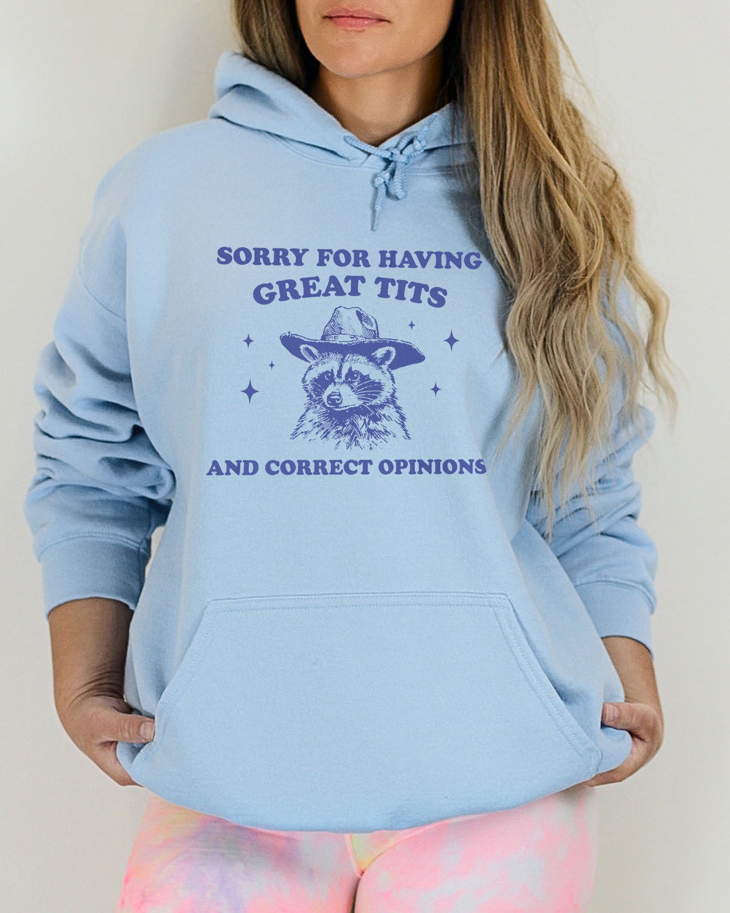 Woman wearing a light blue hoodie with a funny retro cowboy raccoon graphic and the phrase 'Sorry For Having Great Tits And Correct Opinions'.