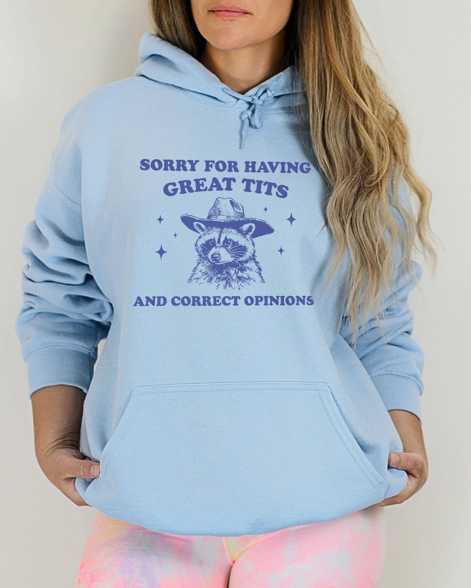 Woman wearing a light blue hoodie with a funny retro cowboy raccoon graphic and the phrase 'Sorry For Having Great Tits And Correct Opinions'.