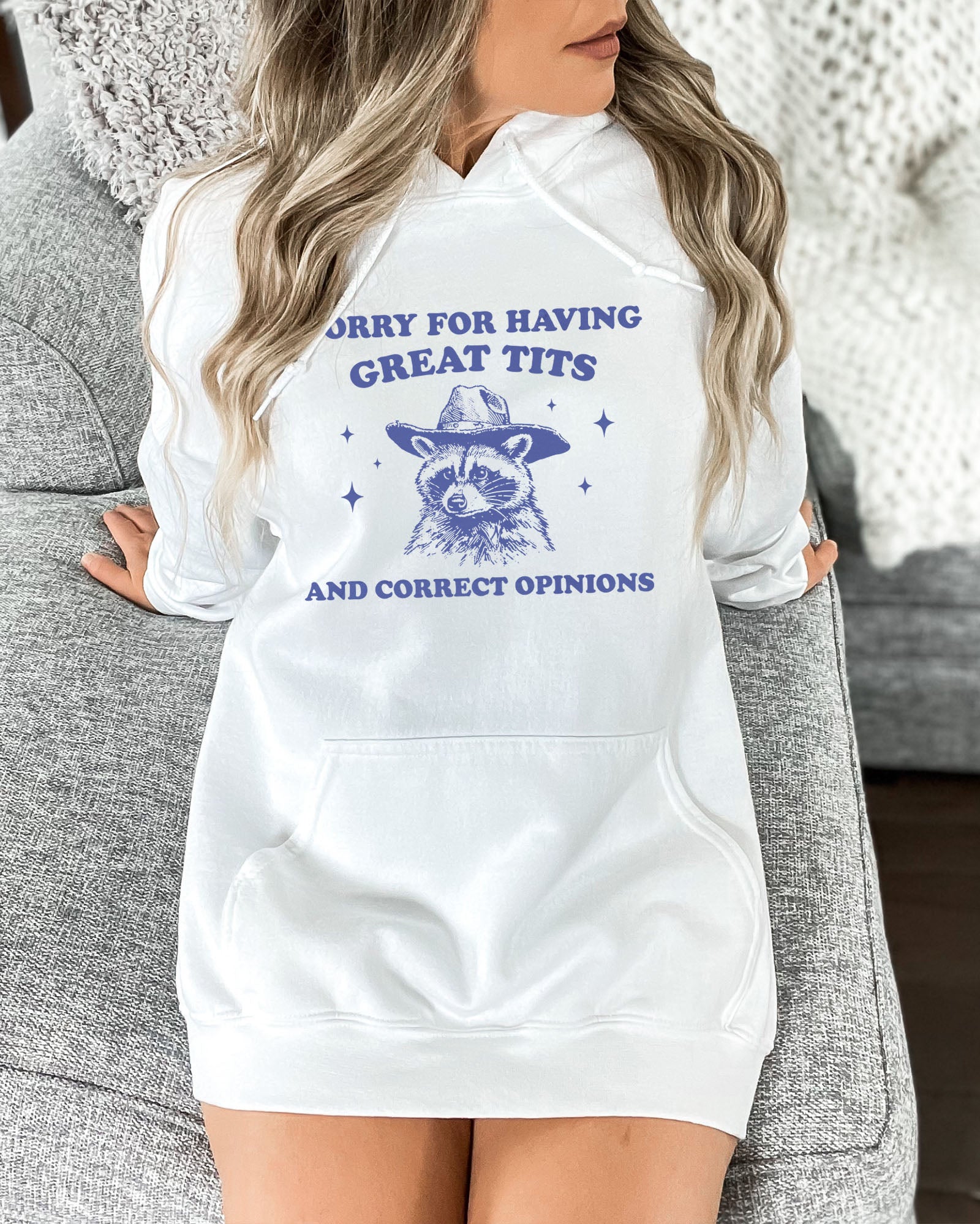 Woman wearing a white hoodie with a funny retro cowboy raccoon graphic and the phrase 'Sorry For Having Great Tits And Correct Opinions'.