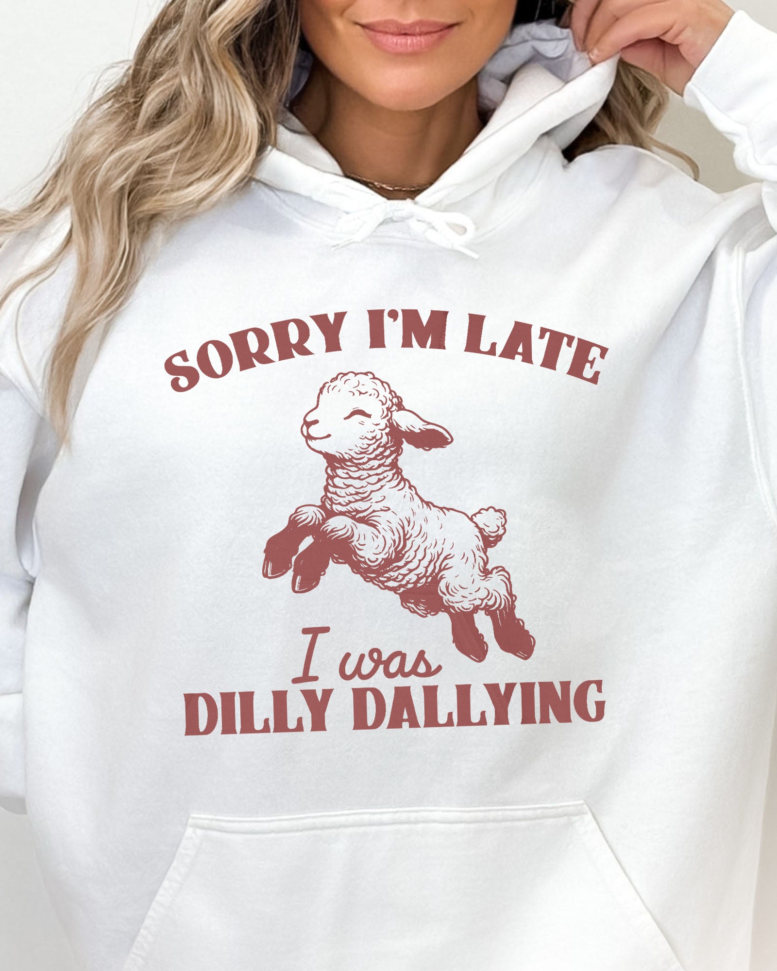 Close-up of a woman wearing a white hoodie featuring a funny retro-style baby sheep graphic and the quote 'Sorry I'm Late, I Was Dilly Dallying '.