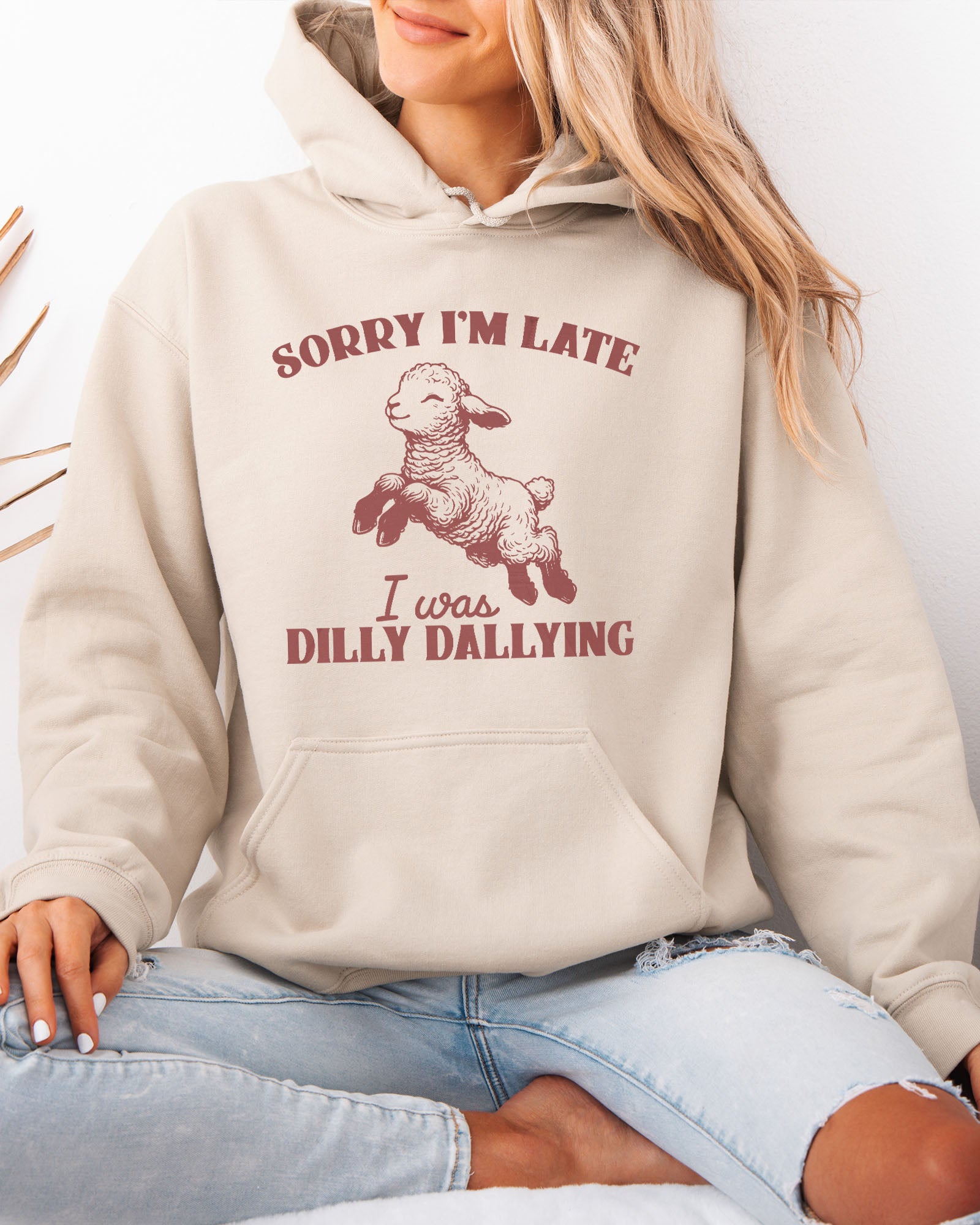 Woman wearing a sand beige hoodie featuring a funny retro-style baby sheep graphic and the quote 'Sorry I'm Late, I Was Dilly Dallying '.