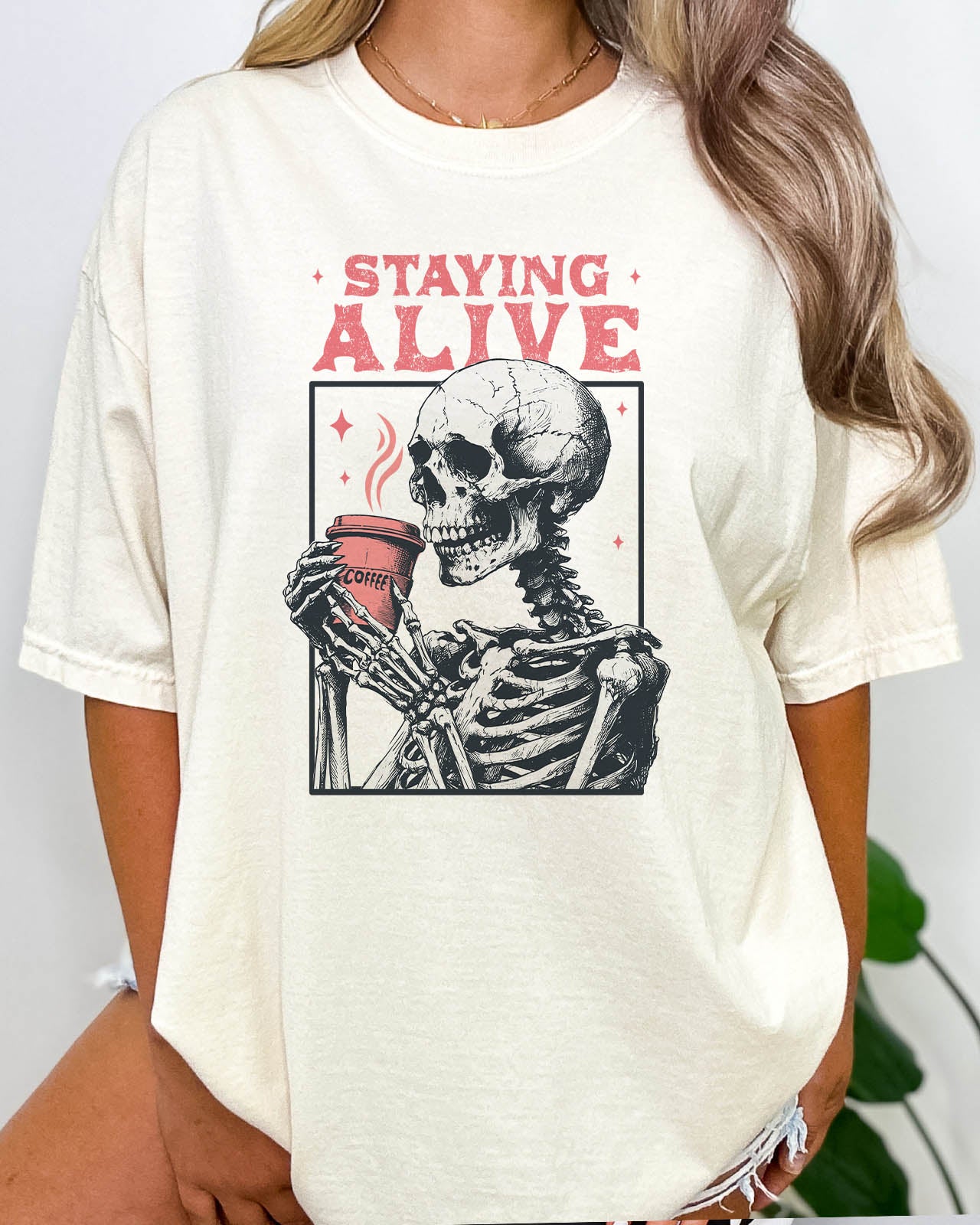 Woman wearing Staying Alive shirt in natural beige color with a funny retro skeleton coffee graphic