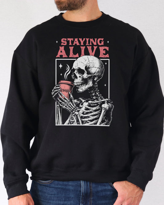 Man wearing a black-colored sweatshirt featuring a retro graphic of a skeleton drinking coffee and the quote 'Staying Alive'.