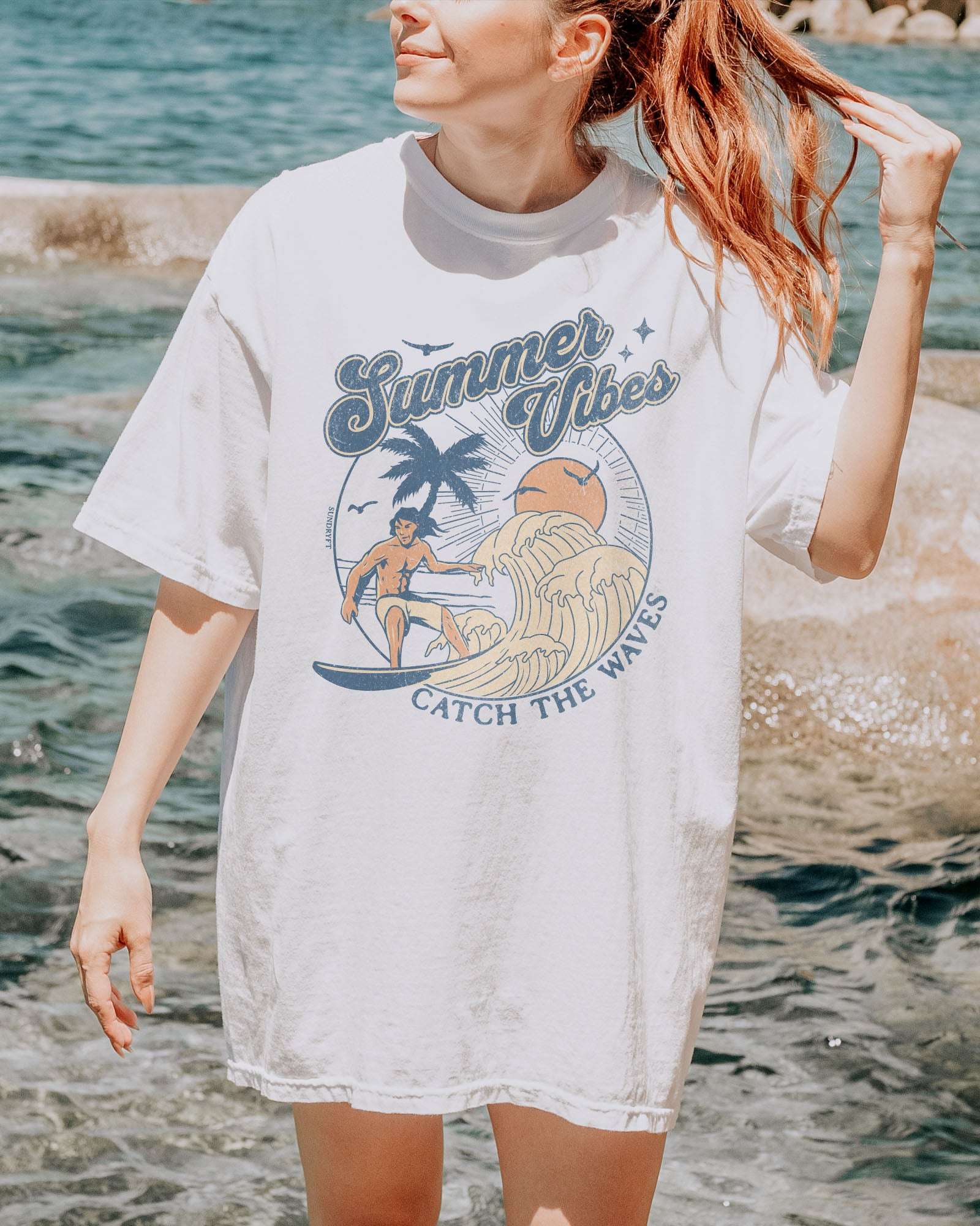 Woman wearing a white 'Summer Vibes – Catch the Waves' surf shirt with a retro graphic of a surfer riding a wave on the beach.