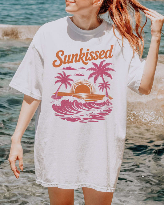 Woman wearing a white-colored 'Sunkissed' shirt with a retro graphic of a sunny beach and ocean waves.