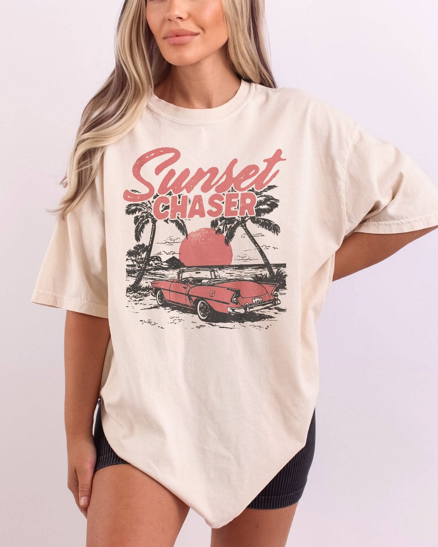 Woman wearing ivory shirt with the quote "Sunset Chaser" and a retro car on a beach scenery graphic.