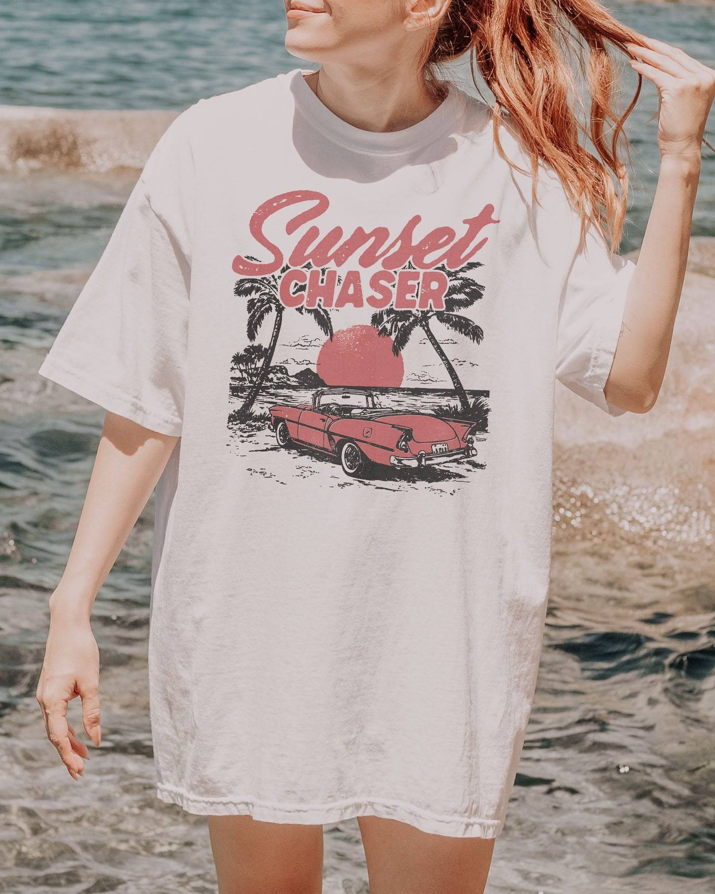 Woman wearing white shirt with the quote "Sunset Chaser" and a retro car on a beach scenery graphic.