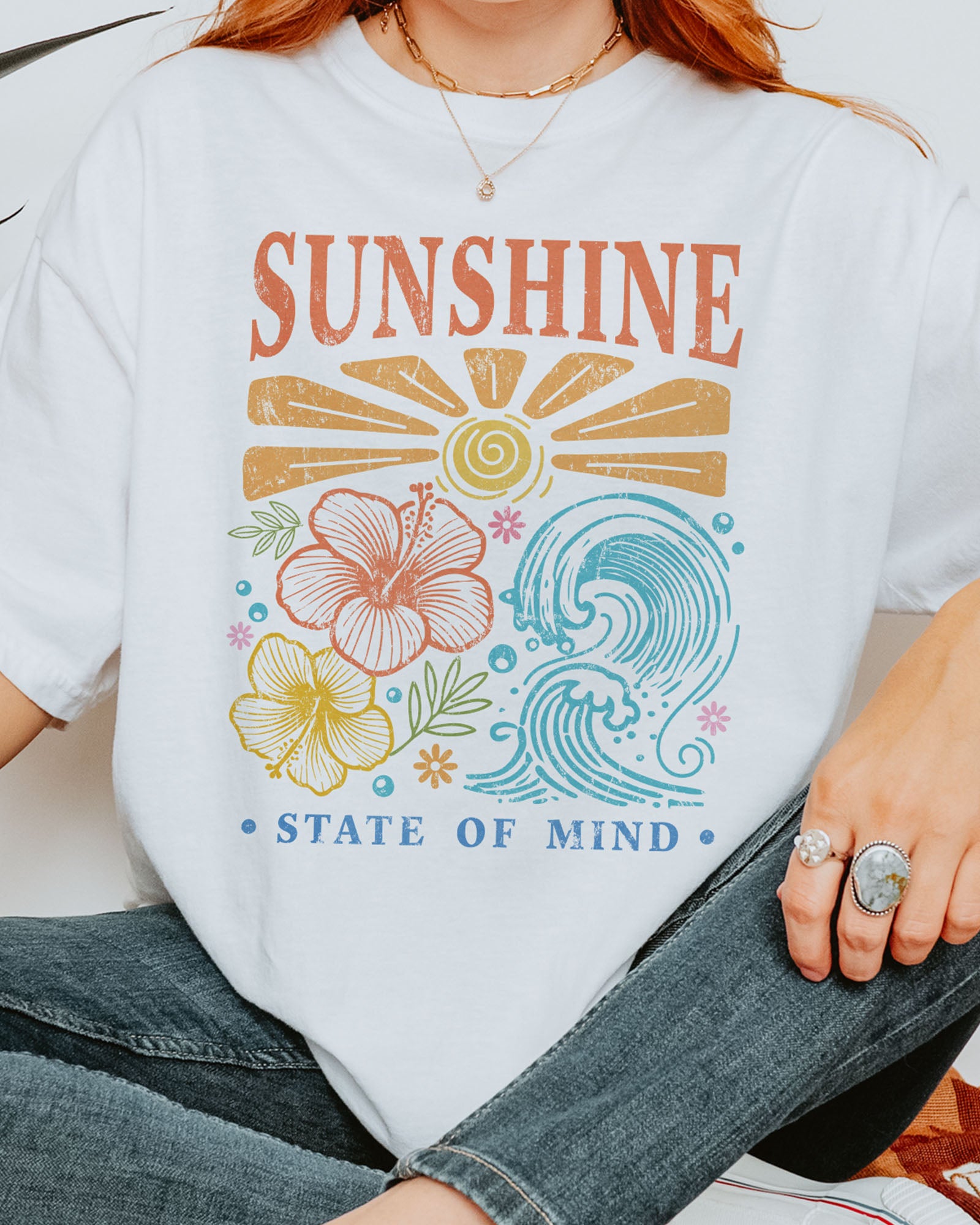 Close-up of woman wearing a white "Sunshine State of Mind" shirt with a retro graphic of outlined flowers, sun, and waves.