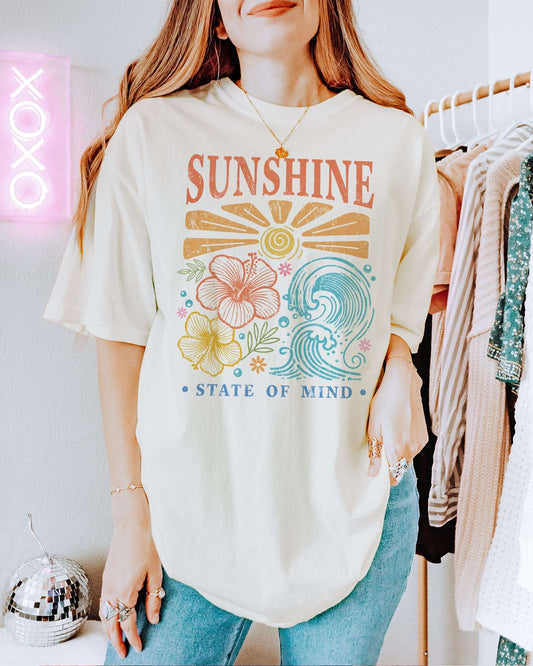 Woman wearing an ivory "Sunshine State of Mind" shirt with a retro graphic of outlined flowers, sun, and waves.