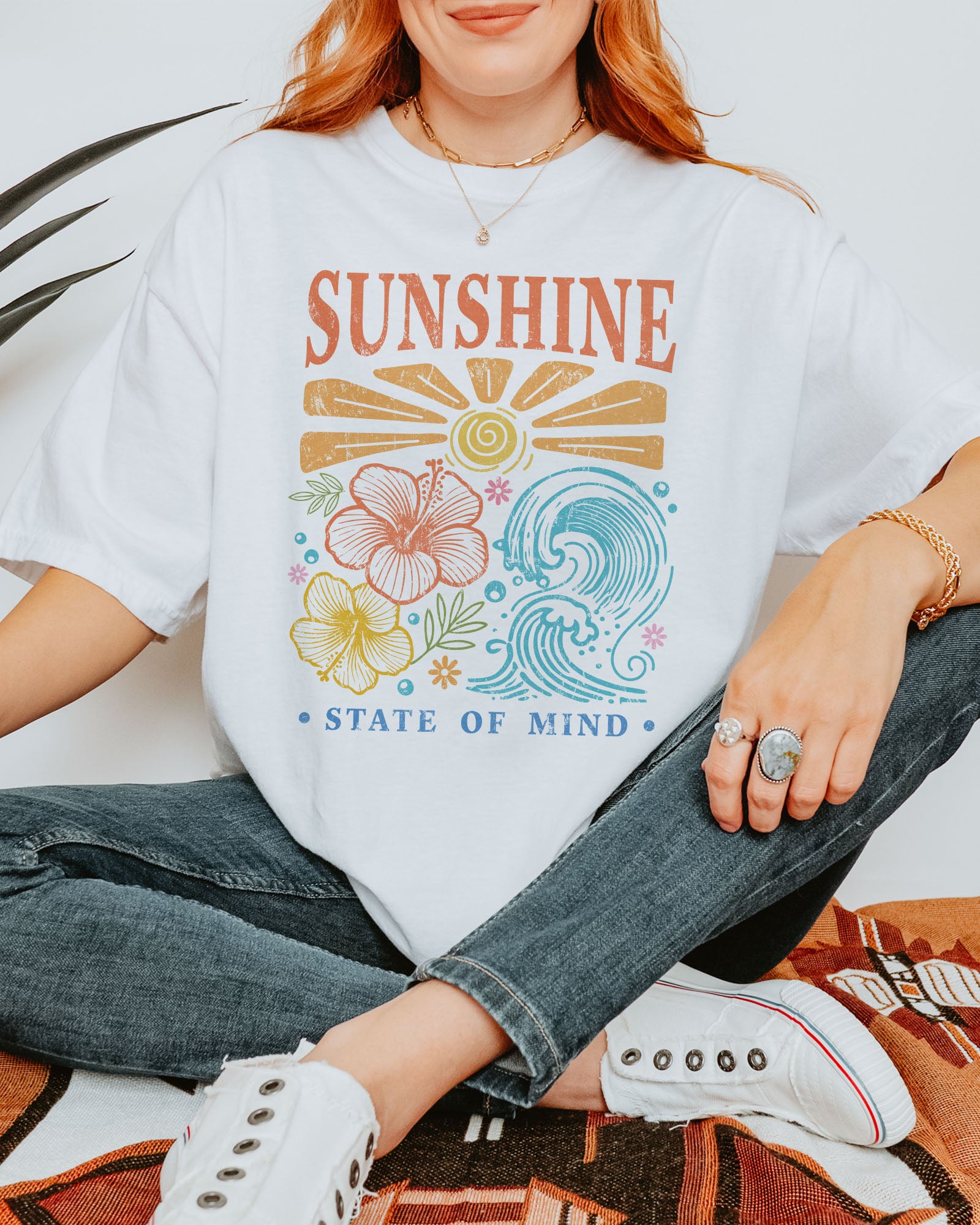 Woman wearing a white "Sunshine State of Mind" shirt with a retro graphic of outlined flowers, sun, and waves.