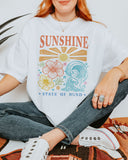Woman wearing a white "Sunshine State of Mind" shirt with a retro graphic of outlined flowers, sun, and waves.
