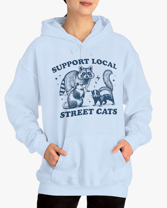 Woman wearing light blue 'Support Local Street Cats' hoodie with a funny raccoon and opossum retro graphic
