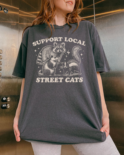Woman wearing a pepper-colored "Support Local Street Cats" shirt with a retro graphic of a raccoon, opossum, and skunk wearing sunglasses.