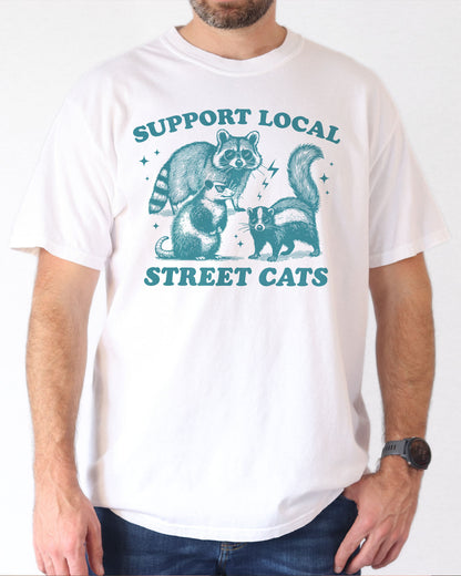Man wearing a white "Support Local Street Cats" shirt with a retro graphic of a raccoon, opossum, and skunk wearing sunglasses.