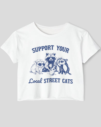 Flat lay of a white 'Support Your Local Street Cats' crop top featuring a funny retro raccoon gang graphic.