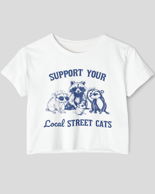 Flat lay of a white 'Support Your Local Street Cats' crop top featuring a funny retro raccoon gang graphic.