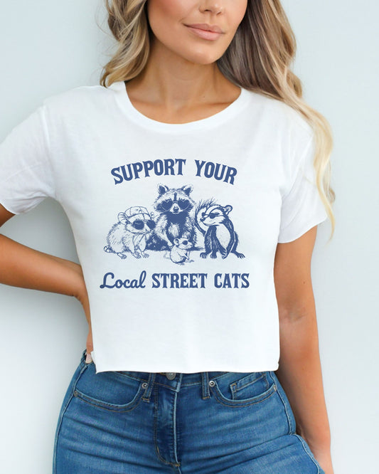 Woman wearing a white 'Support Your Local Street Cats' crop top featuring a funny retro raccoon gang graphic.