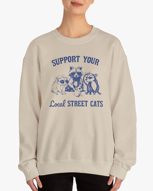 Woman wearing a sand beige sweatshirt with the quote 'Support Your Local Street Cats' and a funny retro raccoon and opossum graphic