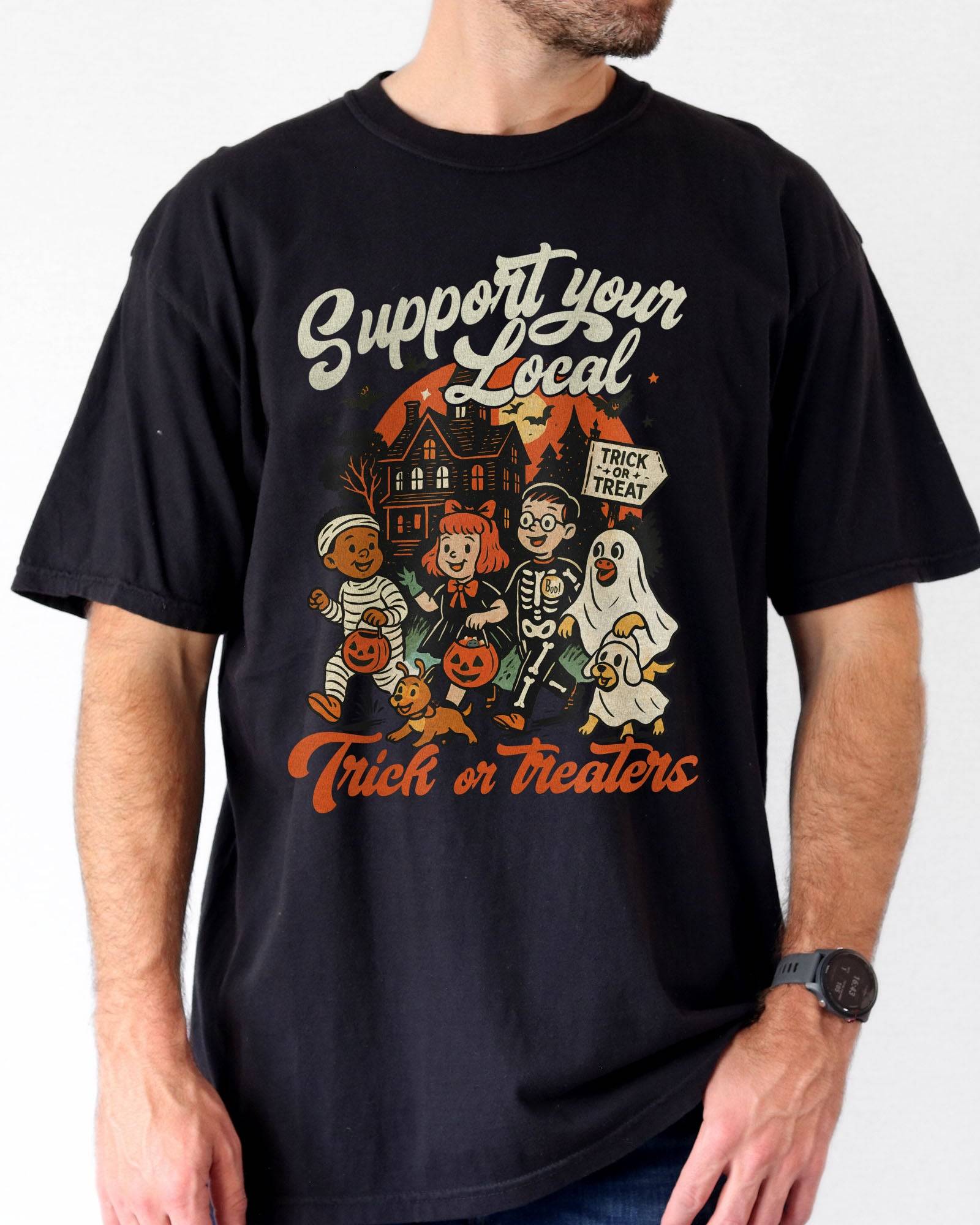 Man wearing a black ‘Support Your Local Trick Or Treaters’ shirt with a retro Halloween graphic of kids trick-or-treating near a haunted house.