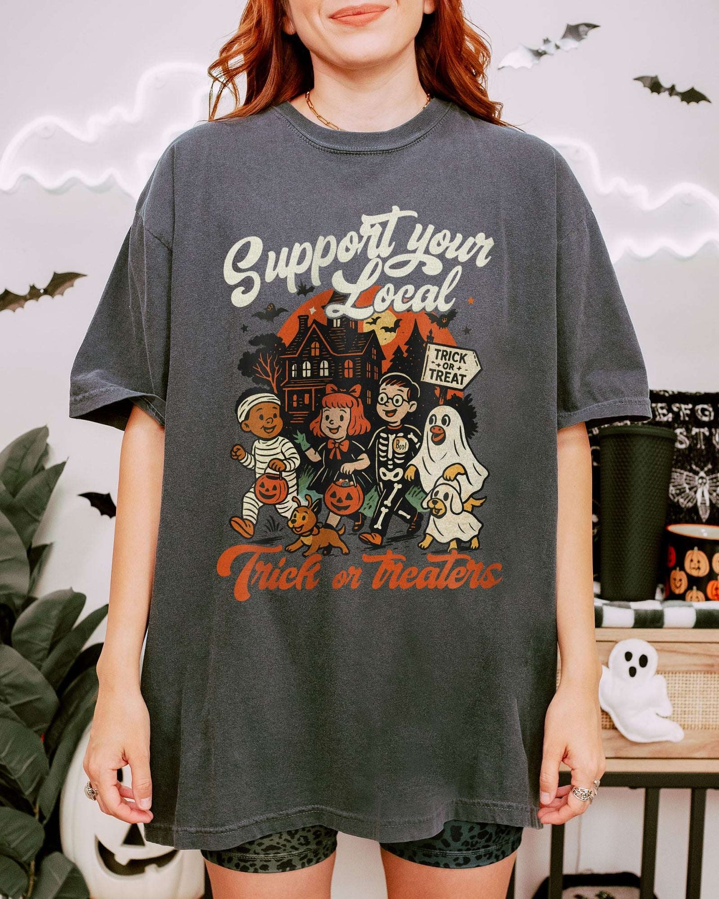 Woman wearing a pepper ‘Support Your Local Trick Or Treaters’ shirt with a retro Halloween graphic of kids trick-or-treating near a haunted house.