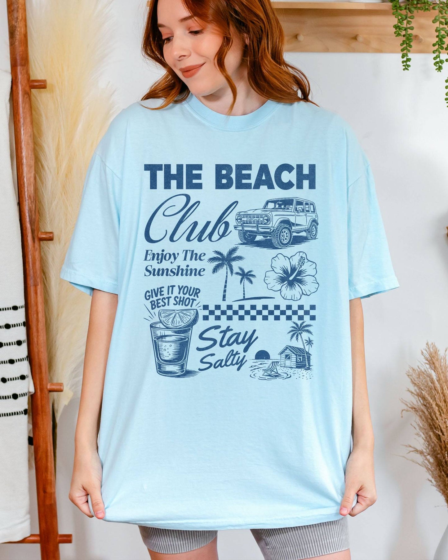 Woman wearing a chambray “The Beach Club” shirt with a retro summer collage of sunshine, beach, and drinks.