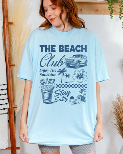 Woman wearing a chambray “The Beach Club” shirt with a retro summer collage of sunshine, beach, and drinks.