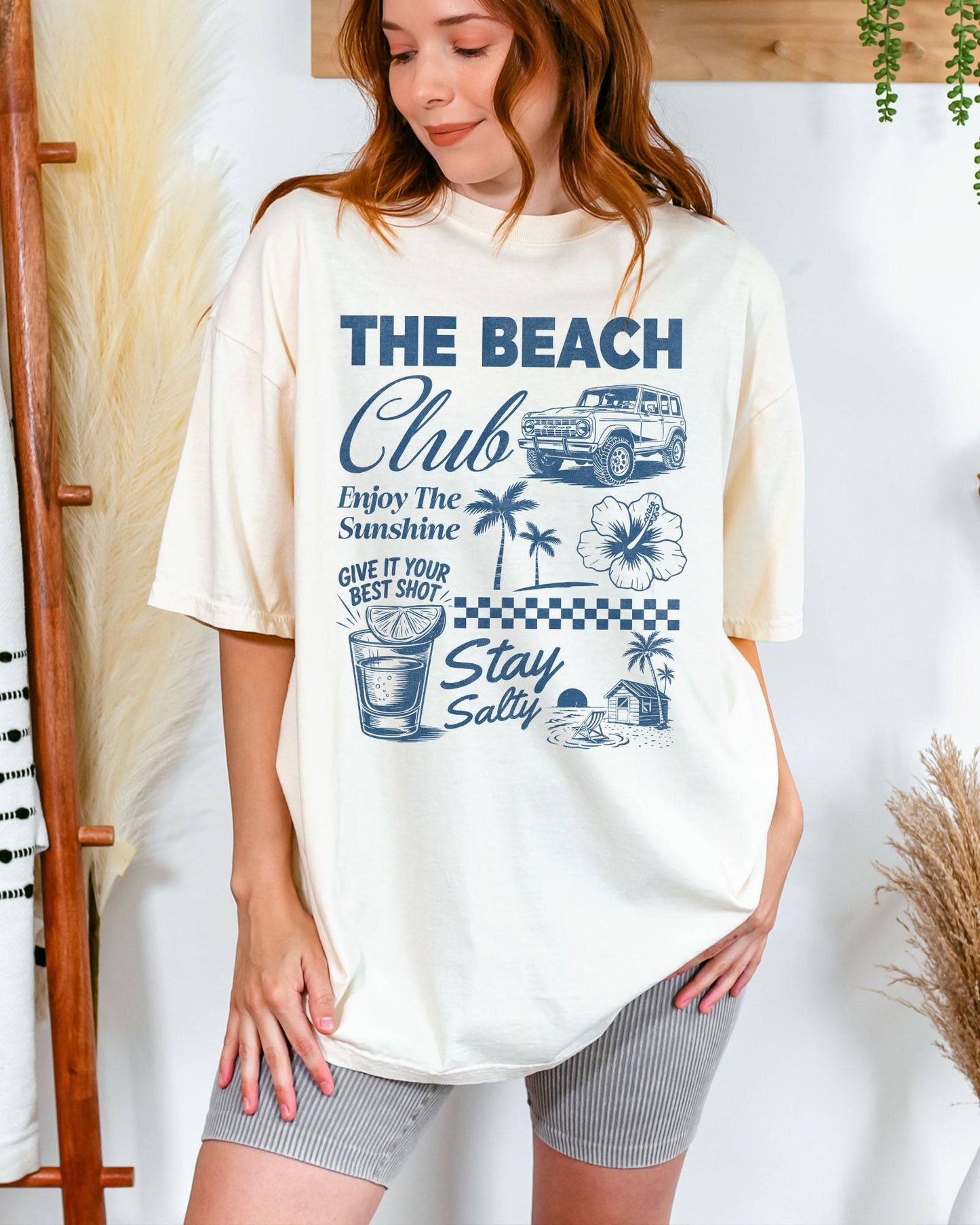 Woman wearing an ivory “The Beach Club” shirt with a retro summer collage of sunshine, beach, and drinks.