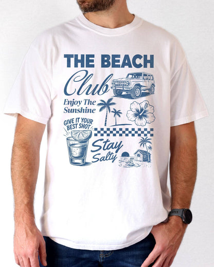 Man wearing a white “The Beach Club” shirt with a retro summer collage of sunshine, beach, and drinks.