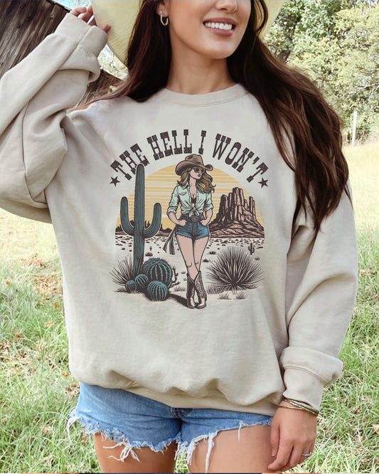 Woman wearing a sand beige-colored sweatshirt with an 80s retro graphic of a cowgirl in a western desert scene and the quote "The Hell I Won’t".