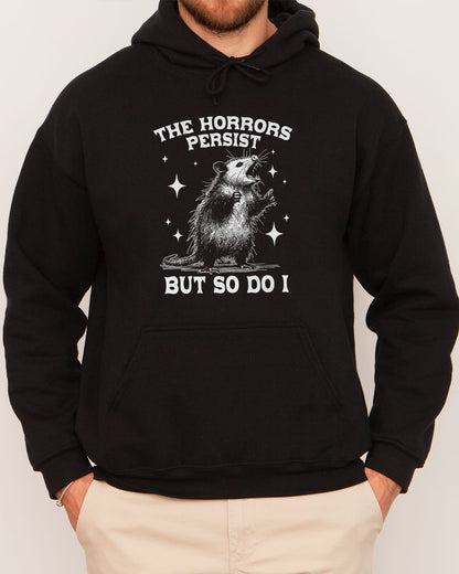 Man wearing a black 'The Horrors Persist But So Do I' hoodie featuring a funny retro yelling opossum graphic.