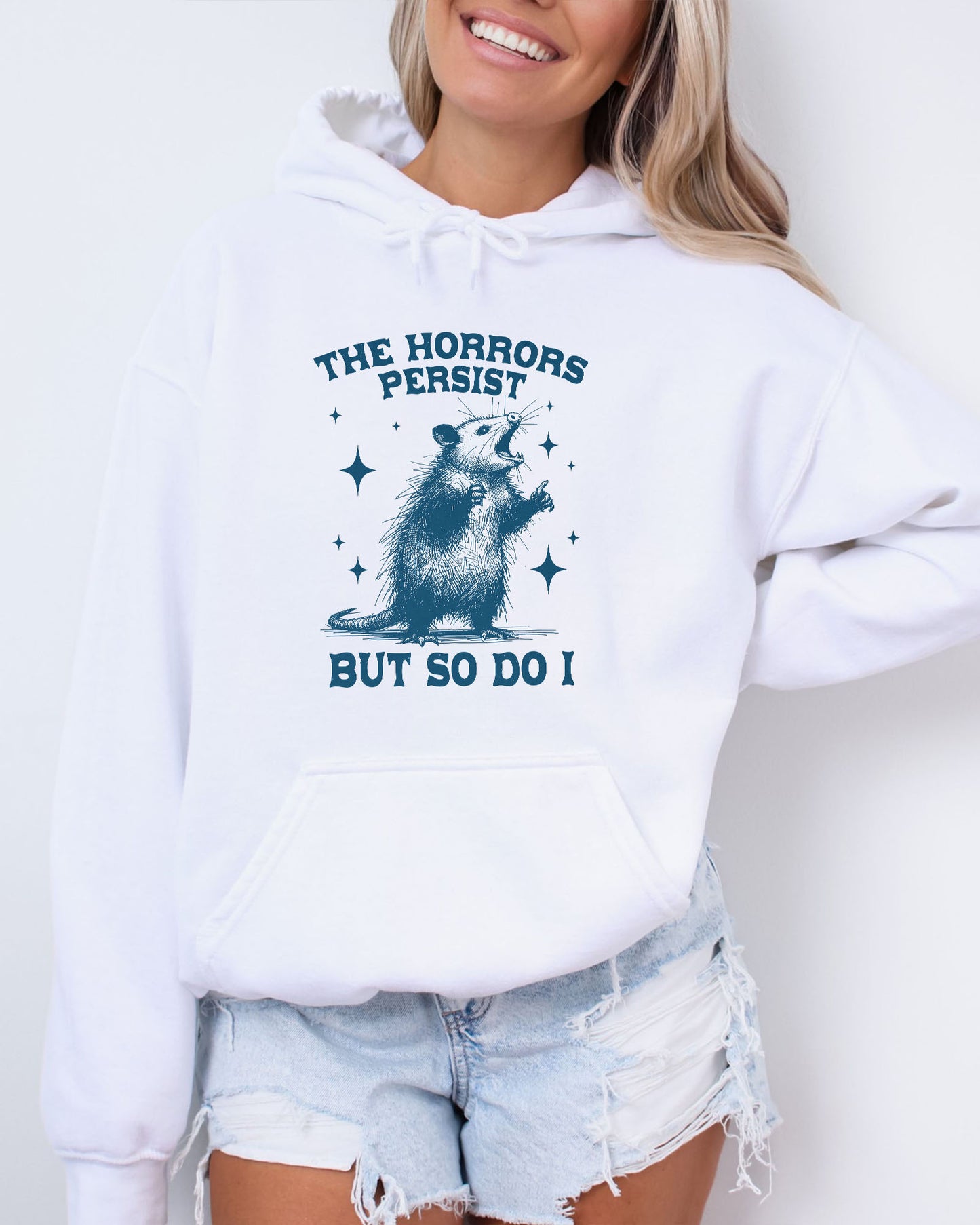 Woman wearing a white 'The Horrors Persist But So Do I' hoodie featuring a funny retro yelling opossum graphic.