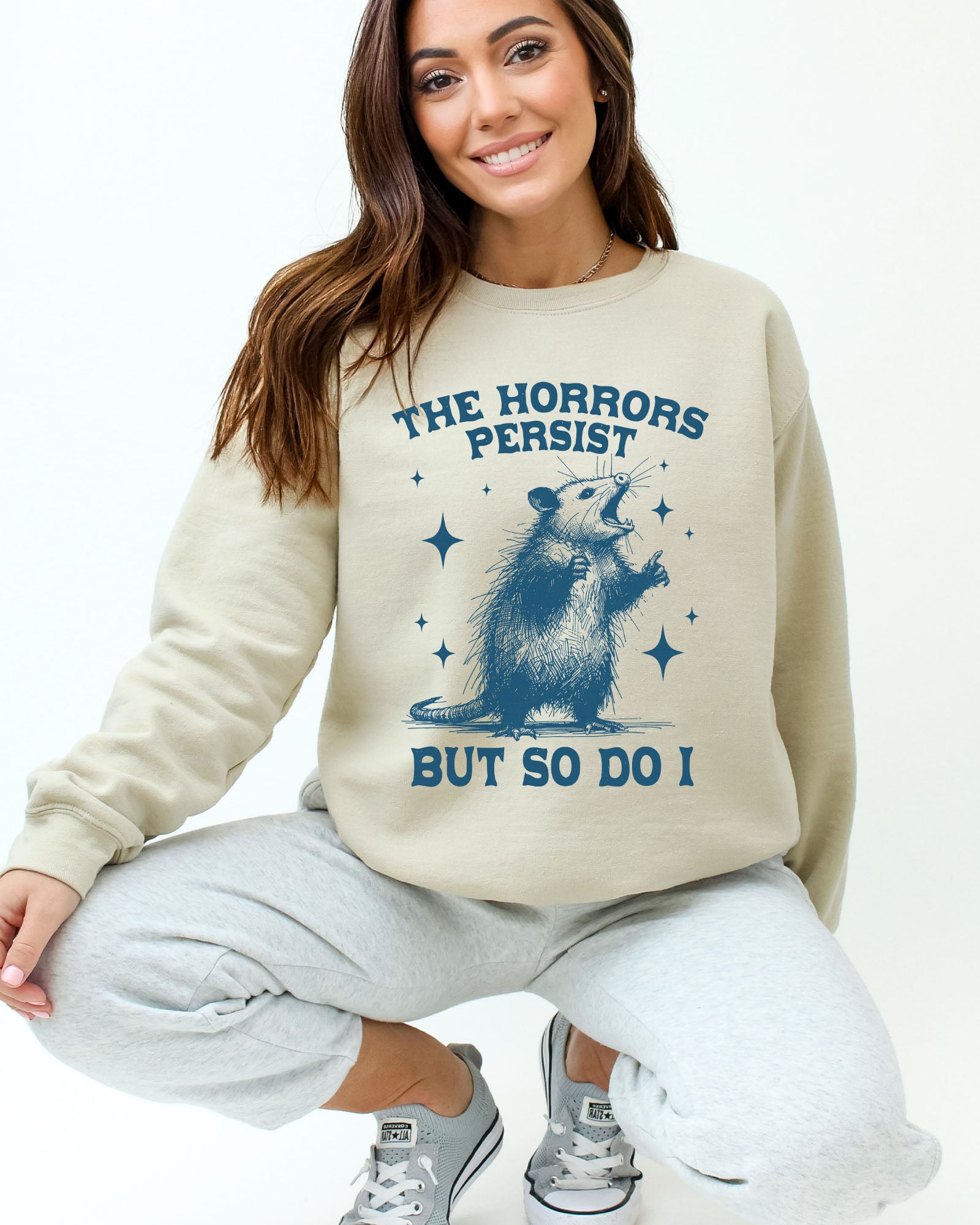 Model wearing sand color sweatshirt with 'The Horrors Persist, But So Do I' graphic design