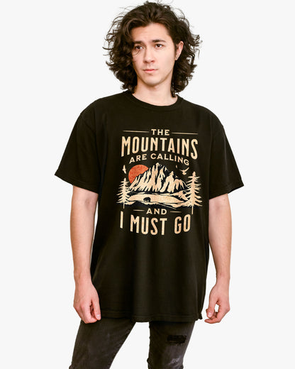 Man wearing black shirt with phrase ‘The Mountains Are Calling And I Must Go’ and retro mountain nature graphic.