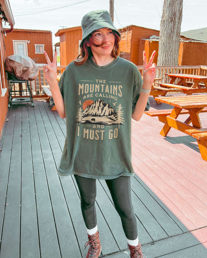 Woman outside wearing moss shirt with phrase ‘The Mountains Are Calling And I Must Go’ and retro mountain nature graphic.